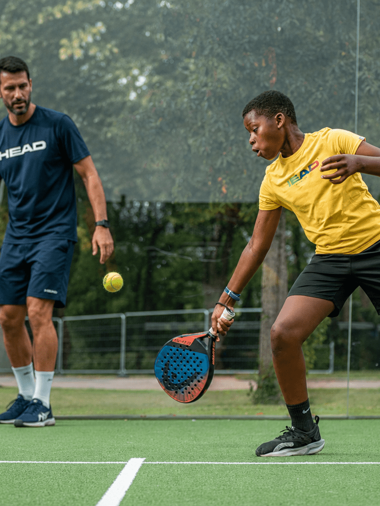 Jeune joueur de padel en plein geste sur un terrain extérieur, concentré sur la balle, accompagné d’un coach adulte en tenue sportive qui l’observe et le guide pendant un cours.